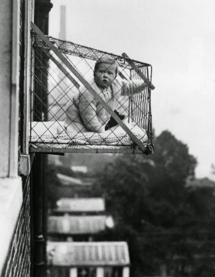 A 1930s window baby cage suspended from the outside of a high-rise apartment window, a real product built to give city babies fresh air